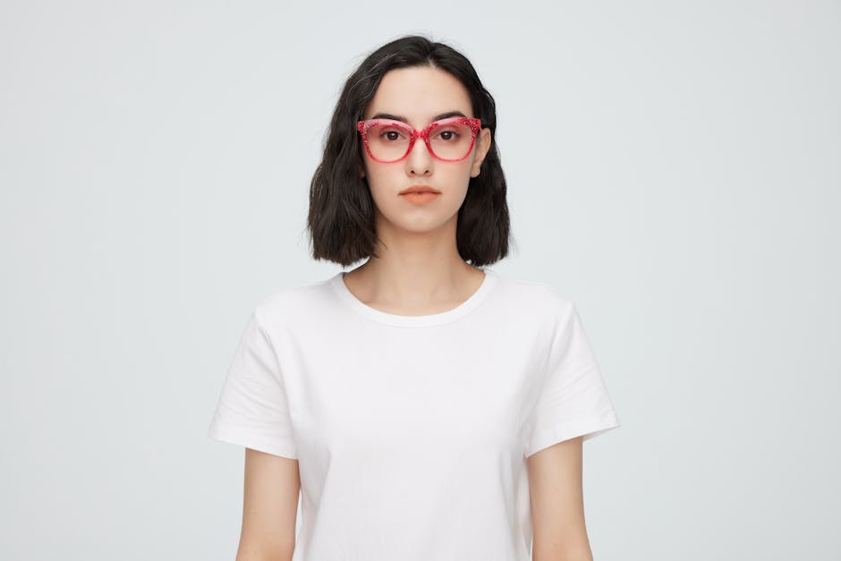 Portrait of a young woman wearing red eyeglasses and a white shirt against a plain background.