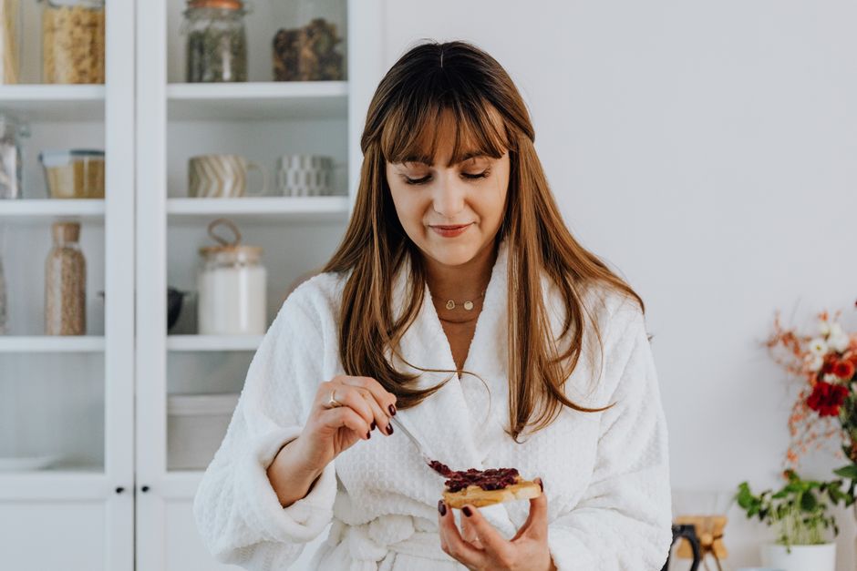 Woman in a bathrobe spreading jam on toast in a bright kitchen.