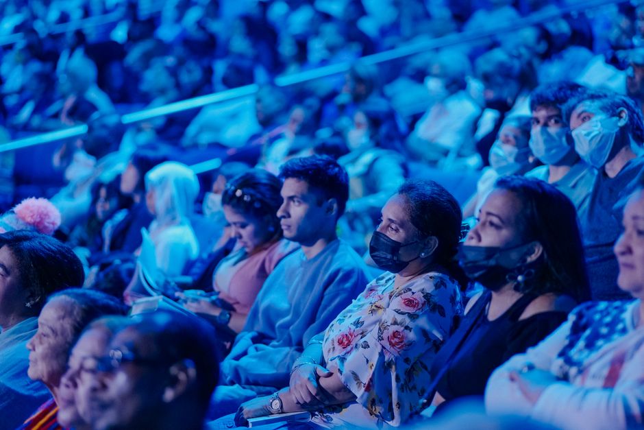A diverse audience in masks attentively watching a concert under blue lights.